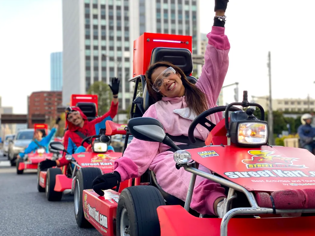 Smiling group of people in red and pink go-karts waving during a street parade in a city setting.